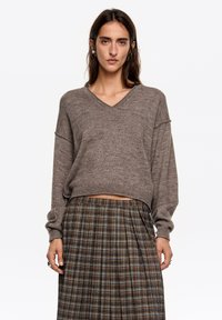 Brązowy sweter na drutach z dekoltem w serek i luźnymi rękawami, w połączeniu z plisowaną spódnicą w kratę z zielonymi, niebieskimi i pomarańczowymi liniami.