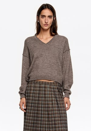 Brązowy sweter na drutach z dekoltem w serek i luźnymi rękawami, w połączeniu z plisowaną spódnicą w kratę z zielonymi, niebieskimi i pomarańczowymi liniami.