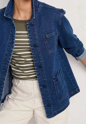 Person iført en blå denimjakke over en olivengrøn og hvid stribet skjorte med cremefarvede bukser.