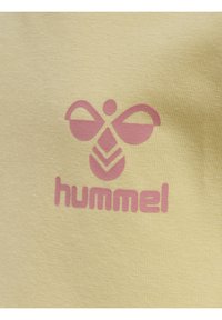 Jasnożółty materiał z różowym logo stylizowanej pszczoły oraz napisem "hummel" poniżej. Gładka tekstura z subtelnym wzorem splotu.