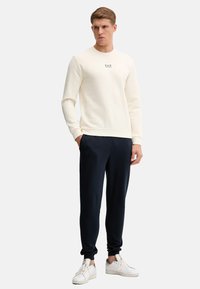 Felpa crema con scollatura rotonda e piccolo logo, abbinata a pantaloni da jogging blu navy e sneakers bianche. Tessuto morbido, vestibilità rilassata e dettagli a costine.