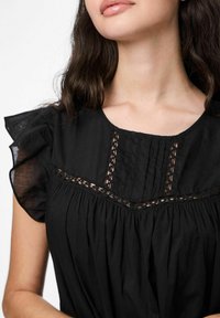 Juoda bluzonė su trumpomis raukšlėtomis rankovėmis, gipiūro akcentais ant kaklo ir suplėtu priekiniu dizainu. Lygaus audinio, laisvo kirpimo.