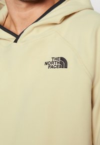Beige fleece hoodie med svart kantat huva och svart broderad "The North Face"-logotyp på bröstet.