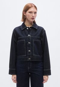 Sötétkék farmervászon rövid dzseki gombos elejjel, nagy mellzsebekkel és fehér varrással díszítve. Csúcsos gallérral és lazán szabott ujjakkal rendelkezik.