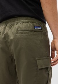 Κοντινό πλάνο ατόμου που φορά ελαφρώς πράσινο παντελόνι cargo με ελαστική μέση και πατρίδα λογότυπο Patagonia στην πίσω τσέπη.