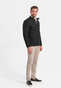 Sort quiltet jakke med høj krave, lynlåslommer og slim fit, parret med lyse beige bukser og sorte glid-sko.