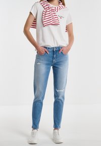 Jeans de mezclilla desgastados de color azul claro con cintura alta, combinados con una camiseta blanca con un pequeño gráfico y una bufanda a rayas rojas y blancas.