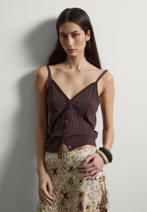 Frau mit langem dunklem Haar, die ein braunes, geknöpftes Tanktop und einen Blumenrock trägt, vor einer schlichten hellen Wand steht und drei Armbänder trägt.