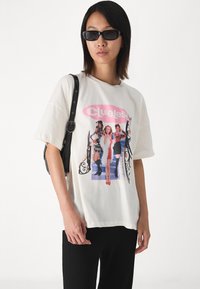 T-shirt blanc surdimensionné avec une impression graphique de trois femmes intitulée "Clueless". Lunettes de soleil carrées noires et petit sac à main noir. Pantalons noirs côtelés.