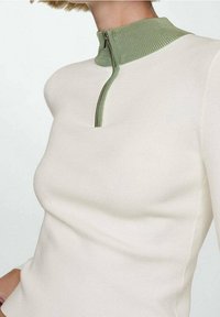 Haut blanc à manches longues avec un col montant côtelé vert et une fermeture éclair partielle. Texture en tricot lisse, coupe ajustée et style minimaliste.