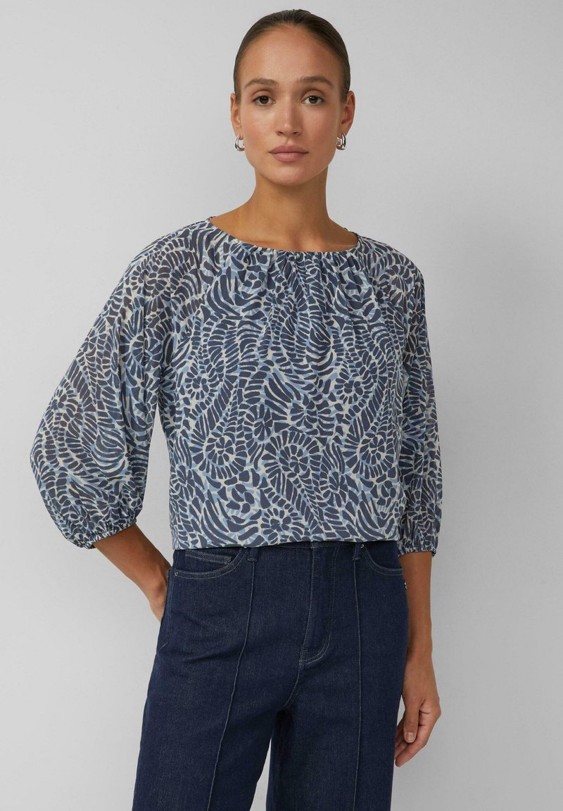 Blau gemusterte Bluse mit Ballonärmeln und gerafftem Ausschnitt, aus fließendem Stoff mit abstraktem botanischem Design. Kombiniert mit dunklem Denim.