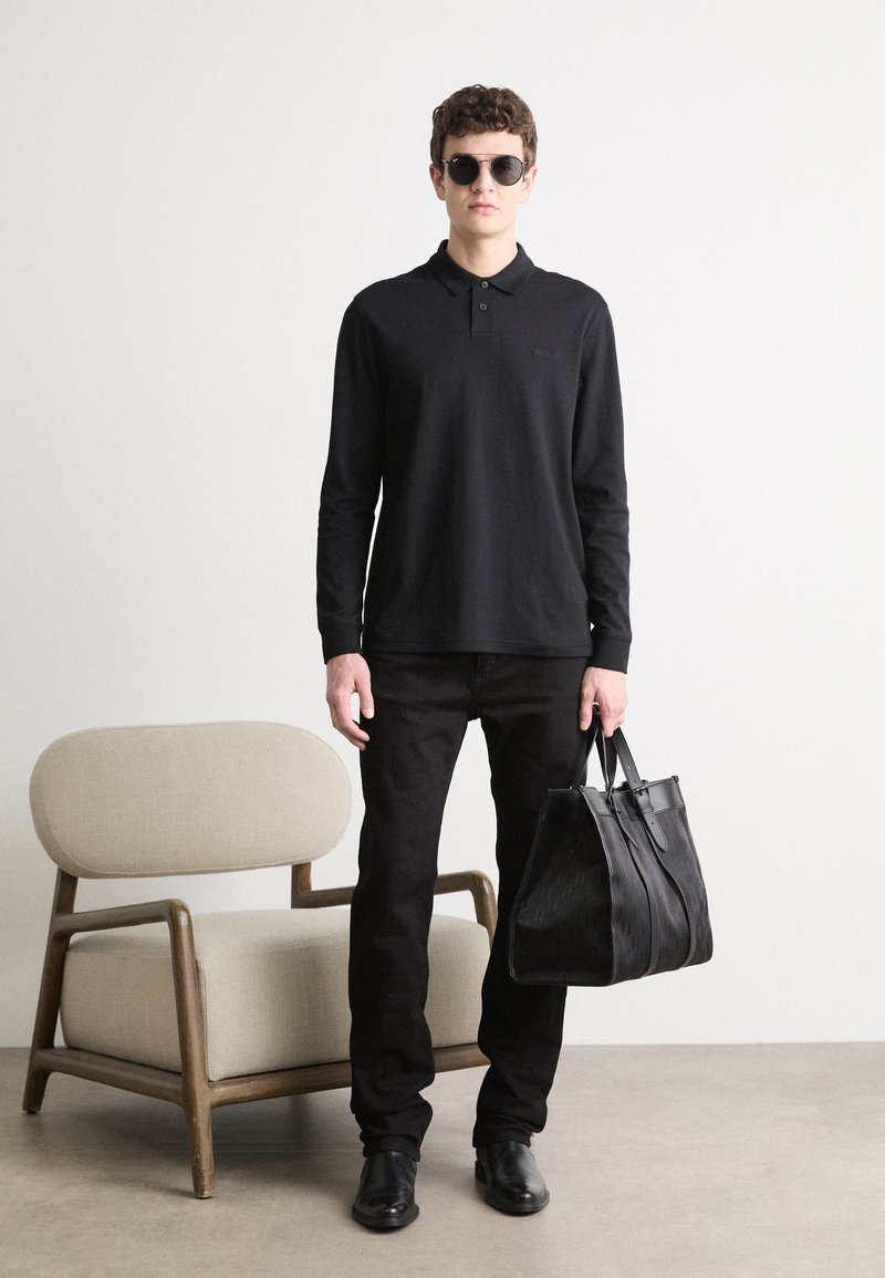 Schwarzes Langarm-Poloshirt mit Kragen, kombiniert mit schwarzen Jeans und Sonnenbrille. Hält eine schwarze strukturierte Tote-Tasche. Vor einem schlichten Hintergrund.