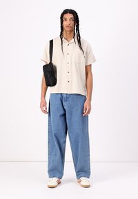 Light beige button-up shirt with textured fabric, paired with loose-fit blue jeans and a small black crossbody bag. White sneakers with red accents.