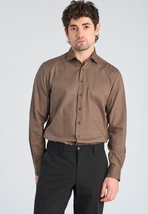 Homme aux cheveux bouclés portant une chemise marron à manches longues boutonnée et un pantalon noir, debout devant un fond clair uni.