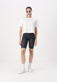 Shorts de vélo avec un imprimé léopard noir associés à un t-shirt court blanc. Le modèle porte des baskets blanches et des lunettes de soleil. Textures de tissu lisses.