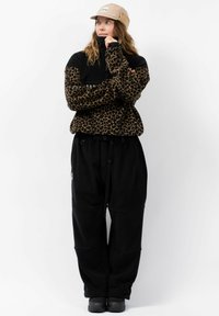 Tijgerprint fleece pullover met zwarte mouwen, gecombineerd met losse zwarte broek. Een beige pet maakt de outfit compleet.