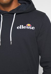 Ciemnogranatowa bluza z kapturem z miękkiego materiału, posiadająca przednią kieszeń kangurkę, białe sznurki oraz wyhaftowane logo „ellesse” z kolorowym motywem słońca.