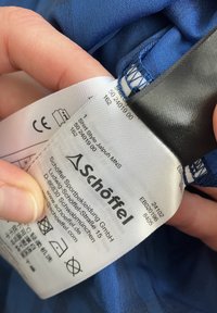 Blaues Hemd mit Pflegeetikett, das das Markenlogo, Waschsymbole und Stilinformationen zeigt. Der Stoff scheint weich und glatt zu sein, mit einer sauberen Nahtverarbeitung.