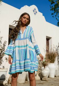 Vestido blanco con patrones geométricos azules, con un escote en V, mangas largas acampanadas y un corte holgado, contra un fondo claro.
