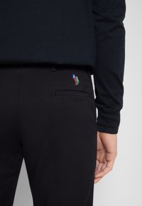 Svarta byxor med en strukturerad yta, med en bakficka och en färgglad broderad logga. En långärmad marinblå tröja är delvist synlig.