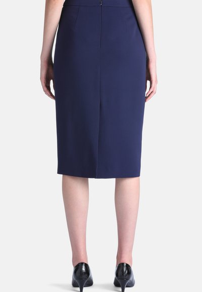 Femme portant une jupe crayon bleu marine longueur genoux et des chaussures noires à talons bas, debout, les mains détendues le long du corps.