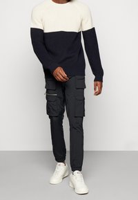 Pull tricoté bleu marine et crème avec un design en blocs de couleur horizontaux, associé à un pantalon cargo noir doté de multiples poches et zips.