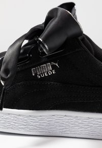 Černé semišové tenisky s bílou gumovou podrážkou, s černou saténovou mašlí jako tkaničkami a kovovým stříbrným nápisem "PUMA SUEDE."