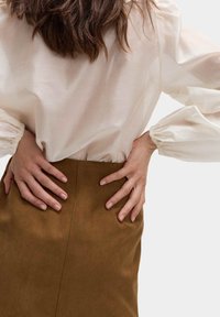 Lichtbeige blouse met opbollende mouwen en elastische manchetten, gecombineerd met een bruine, suède-textuur rok. Handen op de taille geplaatst.