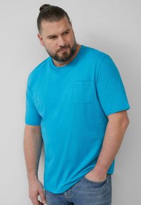 Turquoise kortärm t-shirt van zachte stof met een ronde hals en een borstzak. Gecombineerd met lichte spijkerbroek.