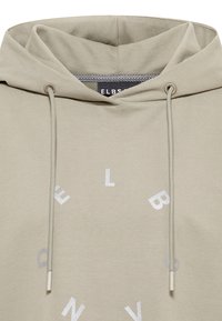 Svetlobéžová mikina s kapucňou a šnúrkami, s jemnou textúrou a nenápadnými logovými detailmi v striebornej farbe na prednej strane.