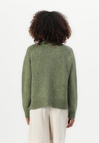 Maglione verde a maglia con colletto alto, spalle scese, orlo a coste e tessuto texturizzato. Vista dal retro, indossato con pantaloni chiari.