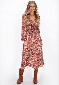Rochie lungă cu mâneci lungi, de culoare roșie, cu decolteu în V, talie strânsă și fustă vaporoasă cu un model ce include accente verzi, asociată cu cizme de gleznă din suede maro.