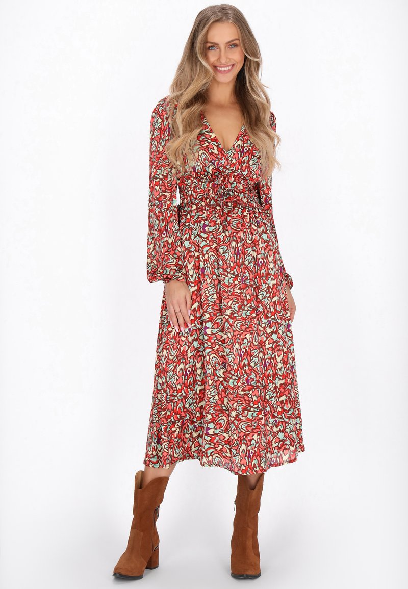 Rochie lungă cu mâneci lungi, de culoare roșie, cu decolteu în V, talie strânsă și fustă vaporoasă cu un model ce include accente verzi, asociată cu cizme de gleznă din suede maro.