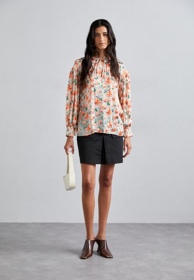 Blusa floral con mangas largas abullonadas en colores pasteles; combinada con una falda negra y zapatos de cuero marrón, sosteniendo un bolso de crema.