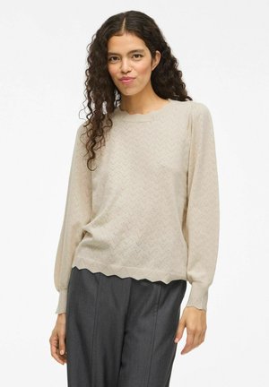 Maglione beige a maglia con motivo a zigzag, orlo scallop e maniche lunghe. Abbinato a pantaloni grigi a sigaretta. Texture liscia.