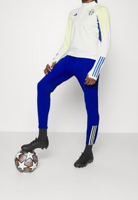 Bílá dlouhá rukávová dres s žlutými a modrými akcenty, s vysokým límcem a emblémem Švédska, doplněná modrými úzkými kalhotami a černými kopačkami.