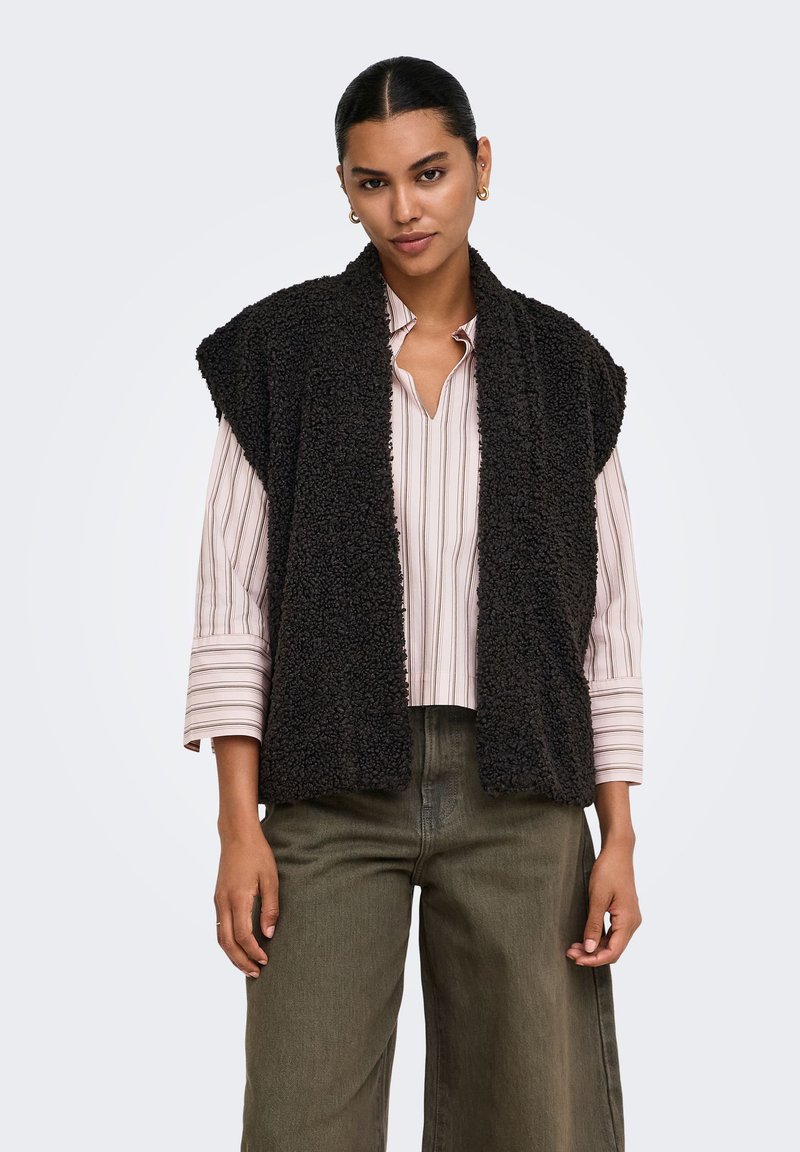 Femme portant un gilet sans manches noir texturé sur une chemise rayée rose clair et un pantalon vert olive, debout devant un fond uni.