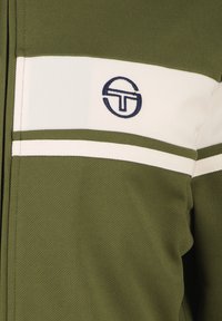 Veste vert olive avec une bande horizontale blanche et un logo brodé en bleu marine. Le tissu a une finition texturée. Détail de fermeture éclair visible.