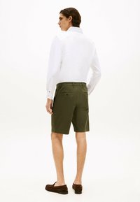 Camisa blanca de manga larga con puños abotonados, combinada con pantalones cortos verde oliva que cuentan con bolsillos traseros y un corte relajado. Zapatos mocasín marrones.