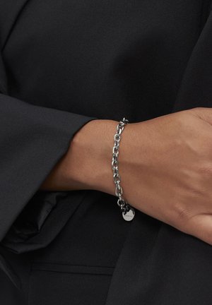 Hånd med sølv kædearmbånd hviler mod sort stof, der viser låsen og lille cirkulær charms detalje.