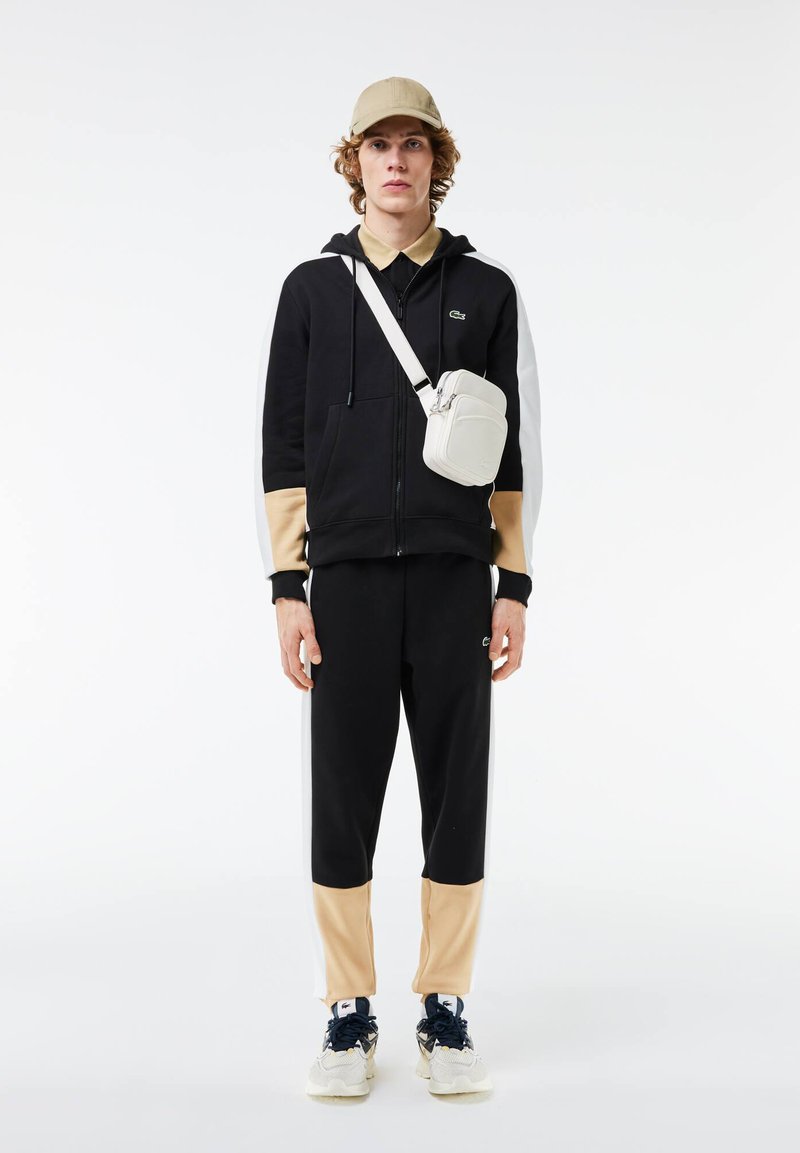 Sweat à capuche noir zippé avec des accents beiges et blancs, pantalon de survêtement noir assorti, casquette beige et sac à bandoulière blanc. Détails de baskets blanches.