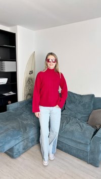 Red knit sweater with high neck and cable pattern, light blue flared jeans, and oversized red sunglasses. Soft, textured blue couch in background.
