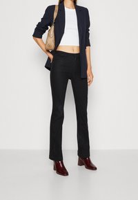 Jeans de campana negros, top corto blanco, blazer azul marino, bolso de mano marrón con estampado y botas de tacón burdeos. Tejido suave y corte a medida.