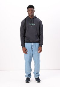 Schwarze Kapuzenjacke mit grafischem Design auf der Brust, kombiniert mit hellblauen Jeans. Das Model trägt schwarze Sneakers und einen schwarzen Schultergurt.