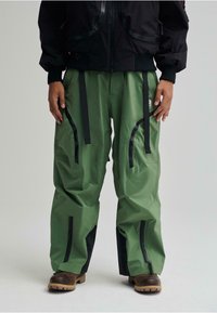 Pantaloni impermeabili verdi con accenti neri, dotati di multiple tasche zippate e spallacci regolabili, caratterizzati da una vestibilità ampia e da un design outdoor.