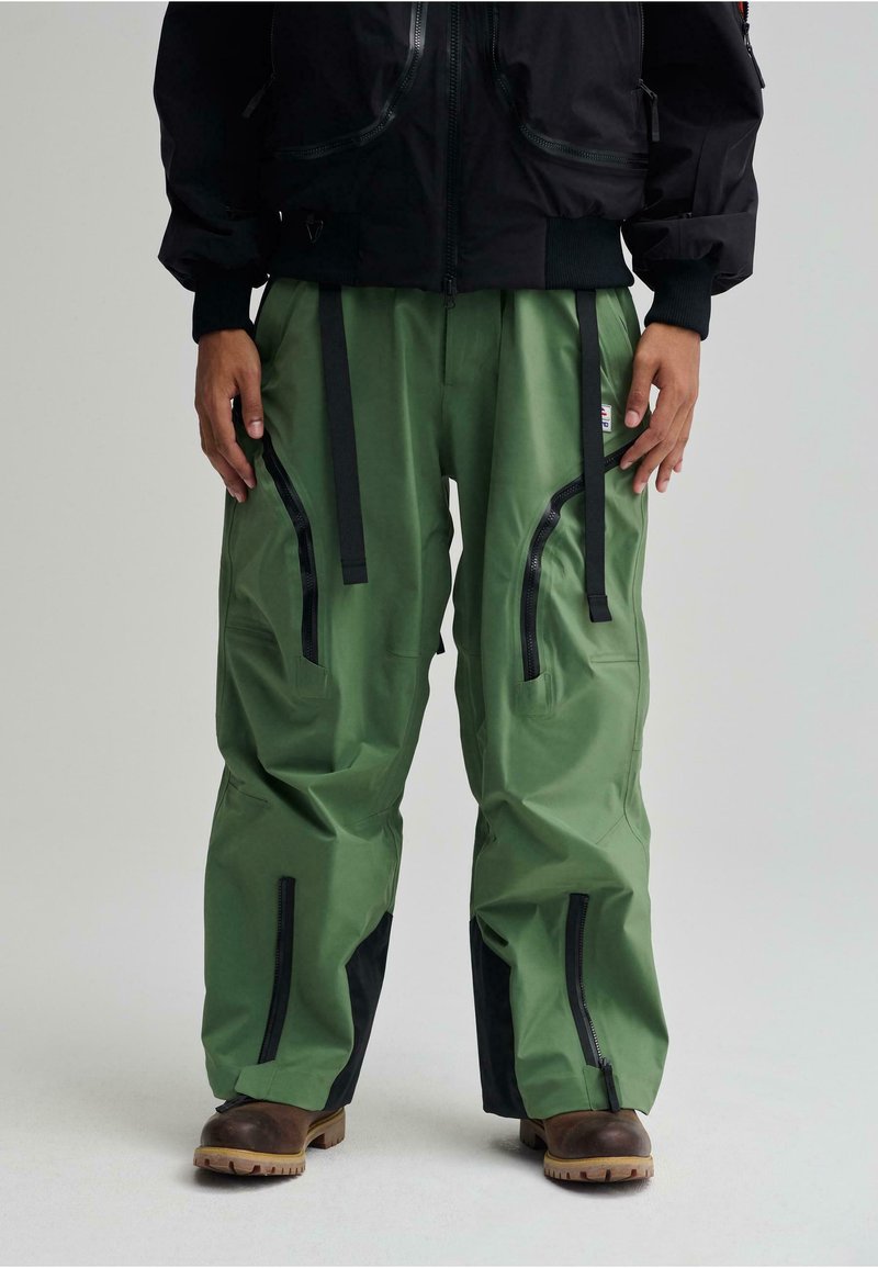 Pantaloni impermeabili verdi con accenti neri, dotati di multiple tasche zippate e spallacci regolabili, caratterizzati da una vestibilità ampia e da un design outdoor.