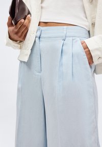 Άτομο που φοράει ανοιχτό μπλε ψηλόμεσα πλισέ παντελόνια, λευκή κοντή μπλούζα και κρεμ σακάκι, κρατώντας ένα καφέ σκούρο τσαντάκι χειρός.