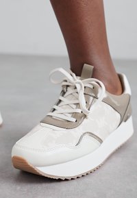 Witte en beige sneaker met een gepatternde bovenkant, texturen materialen, ronde neus en platte zool met een gumrubberen onderzool.