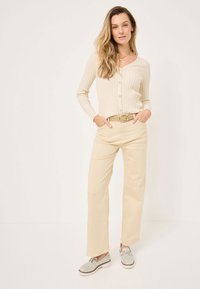 Lichtbeige geribde cardigan met knopen, gecombineerd met crèmekleurige broek met wijde pijpen. Een gouden riem en grijze schoenen met witte zolen maken de outfit af.