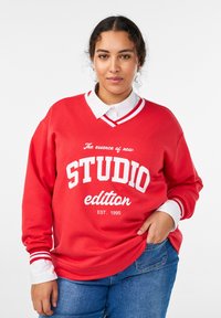 Červená mikina s bielym textom a dizajnom s výstrihom do V. Obsahuje prúžkované biele a červené manžety, položené na košeli s golierom. Neformálny strih.